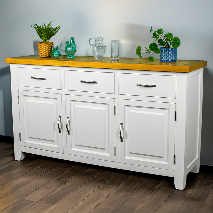 The front of the Felixstowe Large Pine White Buffet. From left to right there are: a potted plant, two blue glass ornaments, a water pitcher, a glass of water and a potted plant on top of the sideboard.