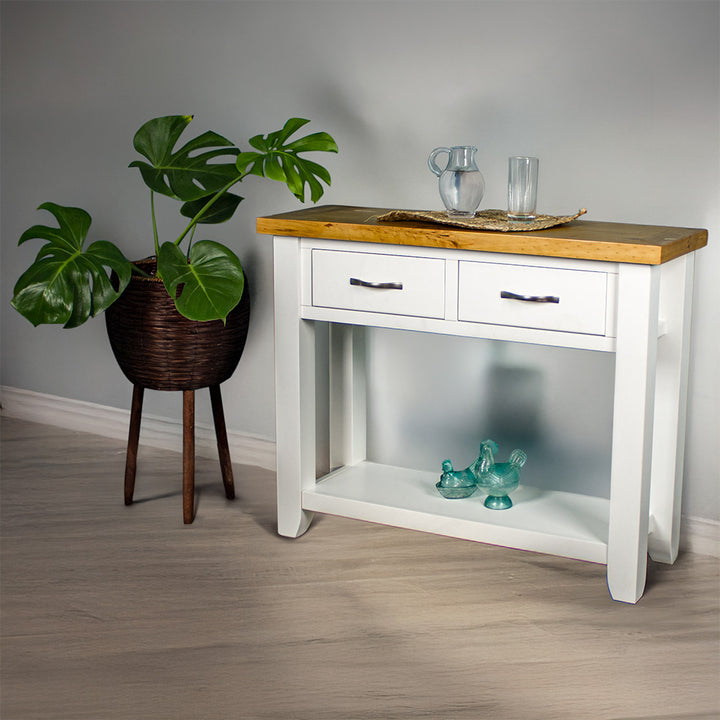 The front of the Felixstowe Large Pine Hall Table. There are two blue glass ornaments on the lower shelf, and a glass pitcher and glass of water on a rattan leaf on top of the table. There is a free standing potted plant next to the hall table.