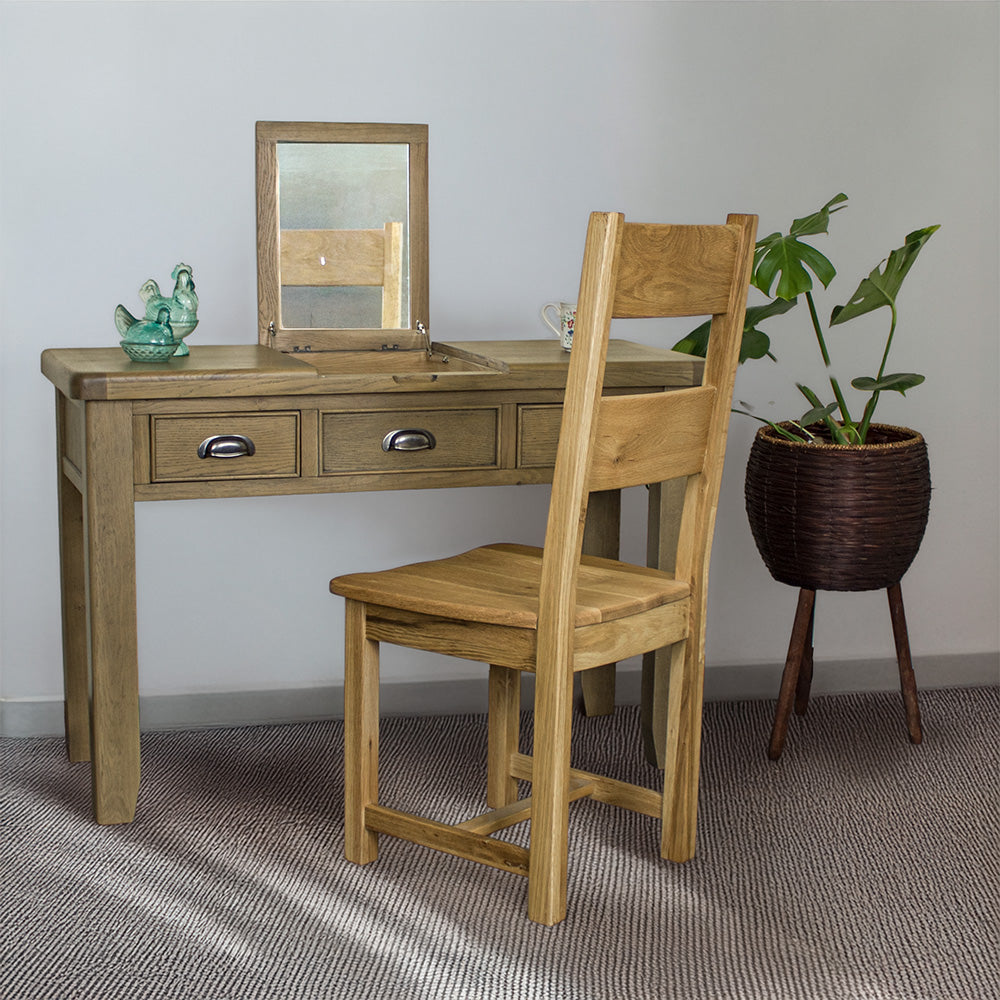 The front of the Houston Oak Dressing Table With Mirror. There is an oak dining chair in front. There are two blue glass ornaments on top of the dressing table and a free standing potted plant to the right of the table.