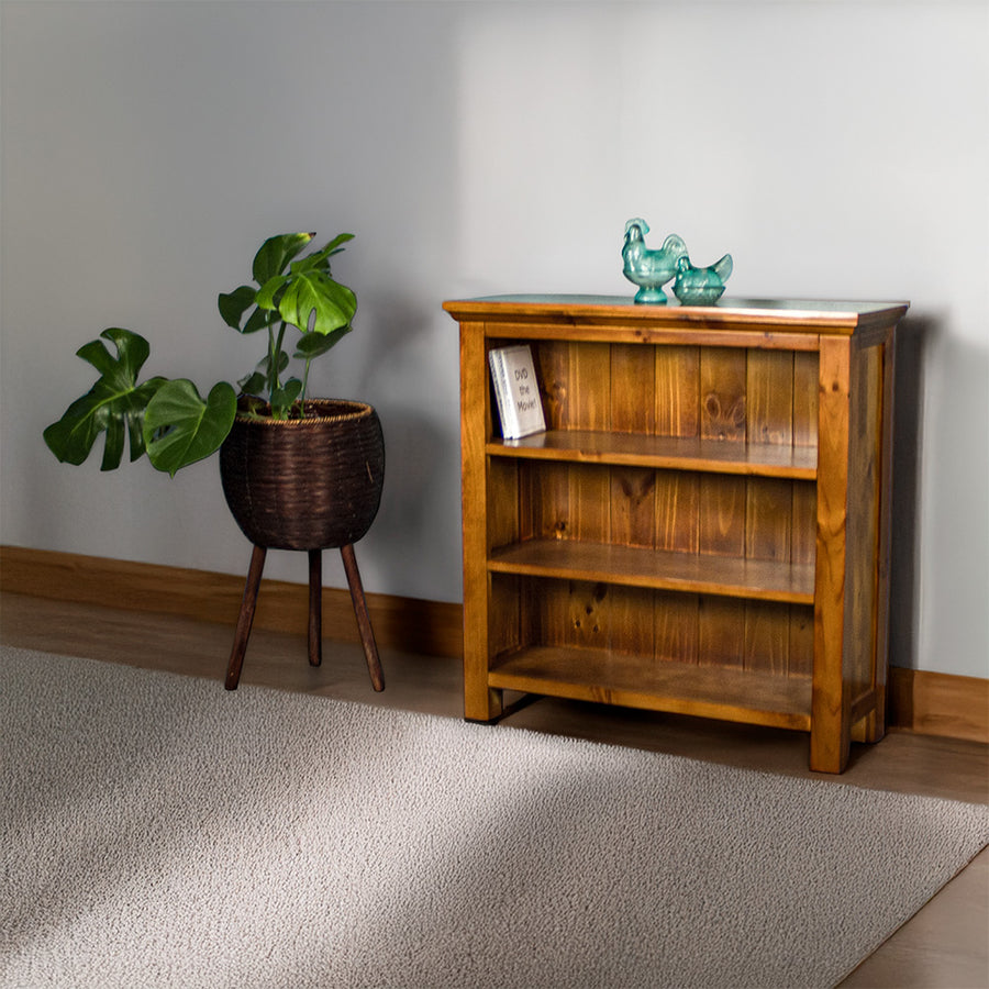 The front of the Montreal Short Pine Bookcase. There are two blue glass ornaments on top and two DVD cases on the top shelf. There is a free standing potted plant next to the bookshelf.