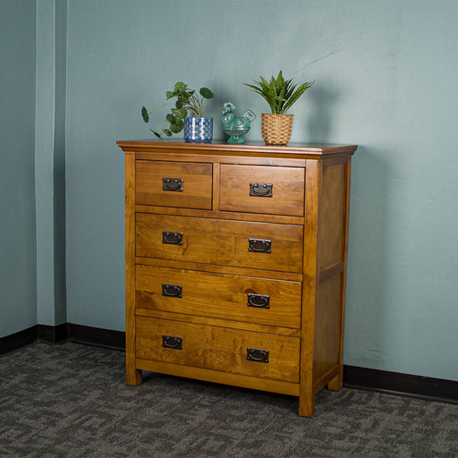 The front of the Montreal Five Drawer Pine Tallboy. There are two potted plants and a blue glass ornament on top.
