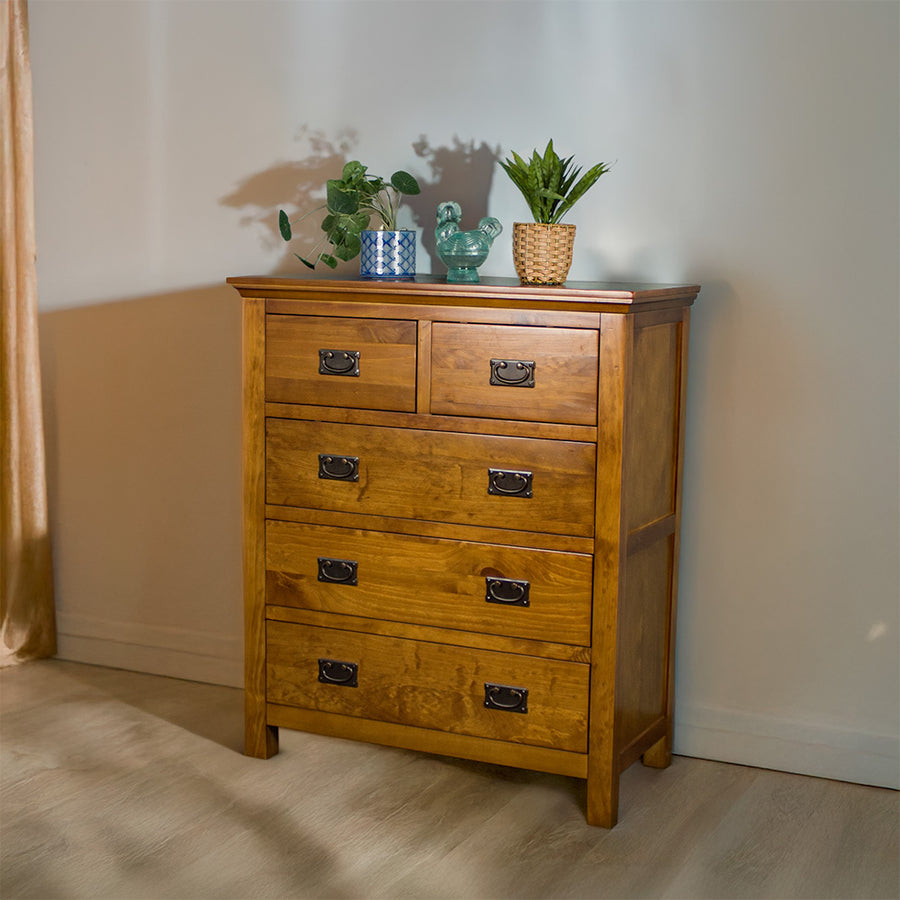 The front of the Montreal Five Drawer Pine Tallboy. There are two potted plants and a blue glass ornament on top.