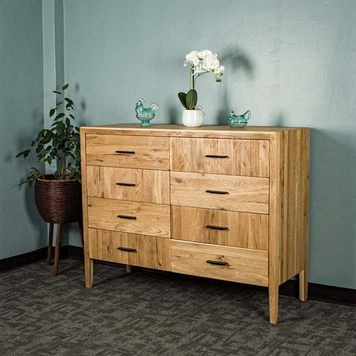 The front of the Ormond 8 Drawer Oak Tallboy. There is a free standing potted plant next to it. There is a small pot of white flowers on top in the middle, with two blue glass ornaments on either side.