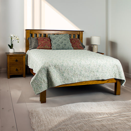 The front of the Trent Queen Size NZ Pine Slat Bed Frame with two Trent Bedside Cabinets next to it. There is a pot of white flowers on the bedside to the left and a lamp on the bedside to the right. The bed is covered in a blue and white sheet, with five pillows on top.
