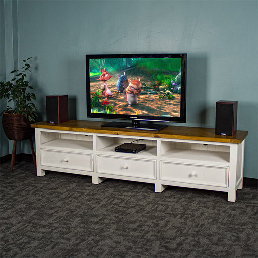 The front of the Tuscan Recycled Pine TV Unit. There is a DVD player in the middle shelf. There is a TV on top with two speakers on either side, and a free standing potted plant next to the TV stand.