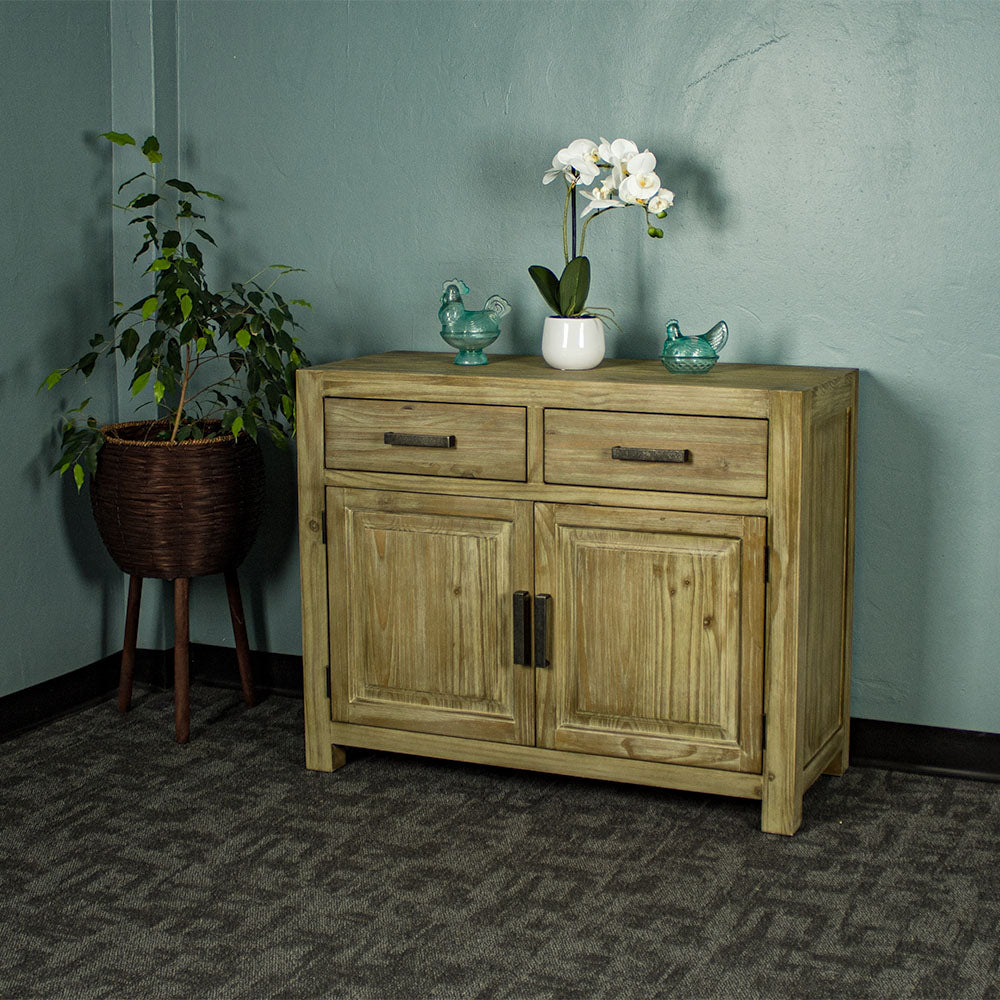 The front of the Vancouver Pine Buffet. There are two blue glass ornaments on top with a pot of white flowers in between. There is a free standing potted plant next to the buffet.