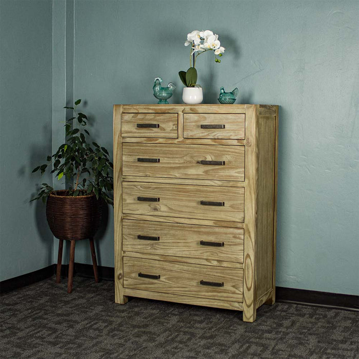 The front of the Vancouver 6 Drawer NZ Pine Tallboy. There are two blue glass ornaments on top with a small pot of white flowers in between. There is a free standing potted plant next to it.