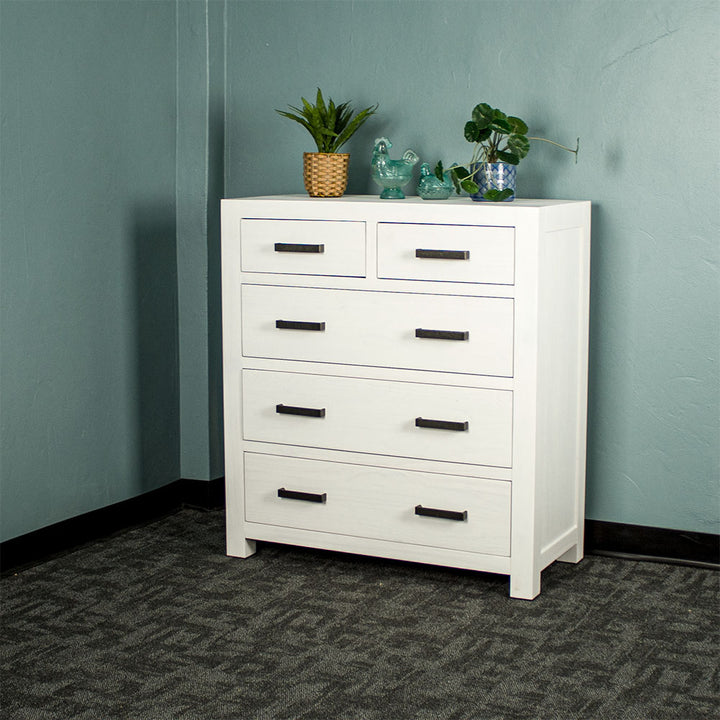 Vancouver Five Drawer White NZ Pine Tallboy front view. There are two potted plants on top and two blue glass ornaments.