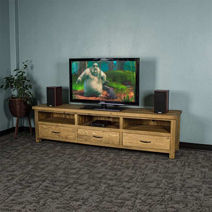 The front of the Ventura Recycled Pine Large TV Unit. There are two speakers on either side of a large TV in the middle on top of the unit. There is a DVD player in the middle shelf. There is a free standing tall potted plant next to the unit.