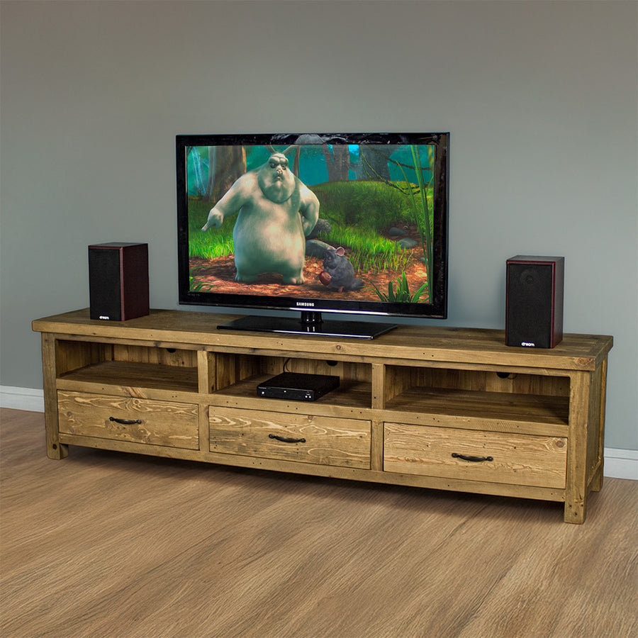 The front of the Ventura Recycled Pine Large TV Unit. There are two speakers on either side of a large TV in the middle on top of the unit. There is a DVD player in the middle shelf. There is a free standing tall potted plant next to the unit.