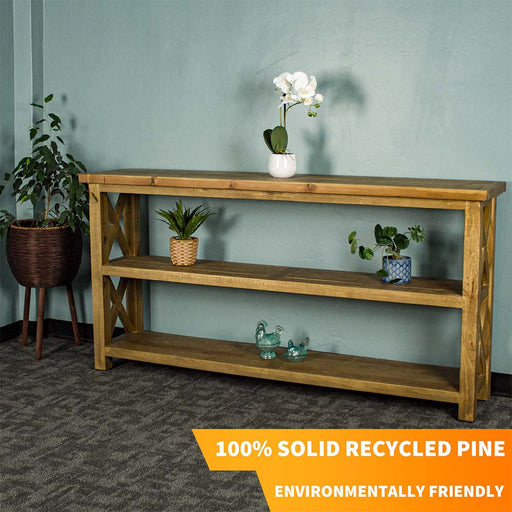 The front of the Ventura Recycled Pine Large Hall Table. There is a free standing potted plant next to it. There is a pot of white flowers on top. There are two equidistant potted plants on the middle shelf. There are two blue glass ornaments on the bottom shelf.