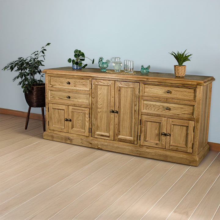 The front of the Versailles Large Oak Buffet. There are two potted plants on either side, and two blue glass ornaments, a pitcher of water next to two glasses on a rattan style leaf coaster in the middle on top.
