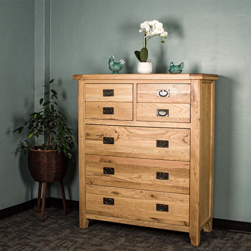 The front of the Vienna 7 Drawer Oak Tallboy. There are two blue glass ornaments on top with a pot of white flowers in between. There is a free standing potted plant next to the unit.