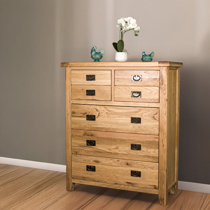 The front of the Vienna 7 Drawer Oak Tallboy. There are two blue glass ornaments on top with a pot of white flowers in between. 