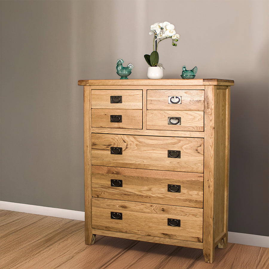 The front of the Vienna 7 Drawer Oak Tallboy. There are two blue glass ornaments on top with a pot of white flowers in between. 