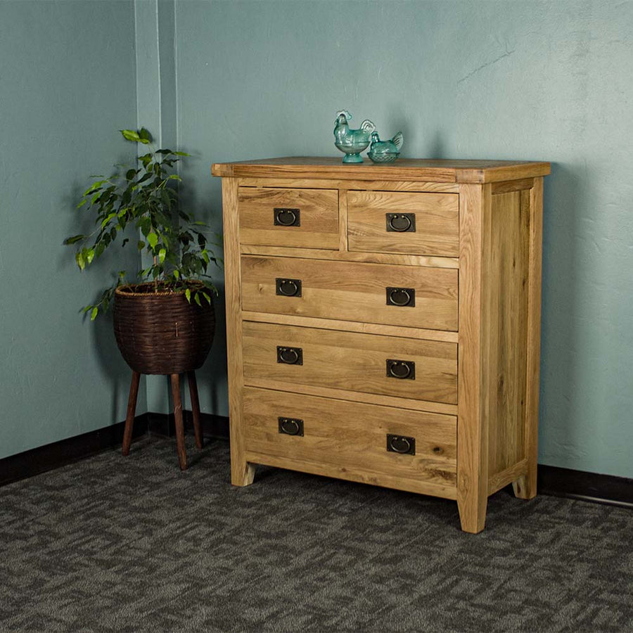 The front of the Yes Five Drawer Oak Chest with two blue glass ornaments on top. There is a free standing potted plant next to the tallboy.