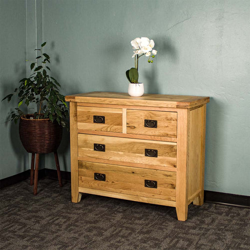 The front of the Yes Four Drawer Oak Lowboy, there is a small pot of white flowers on top and a free standing potted plant next to the lowboy.