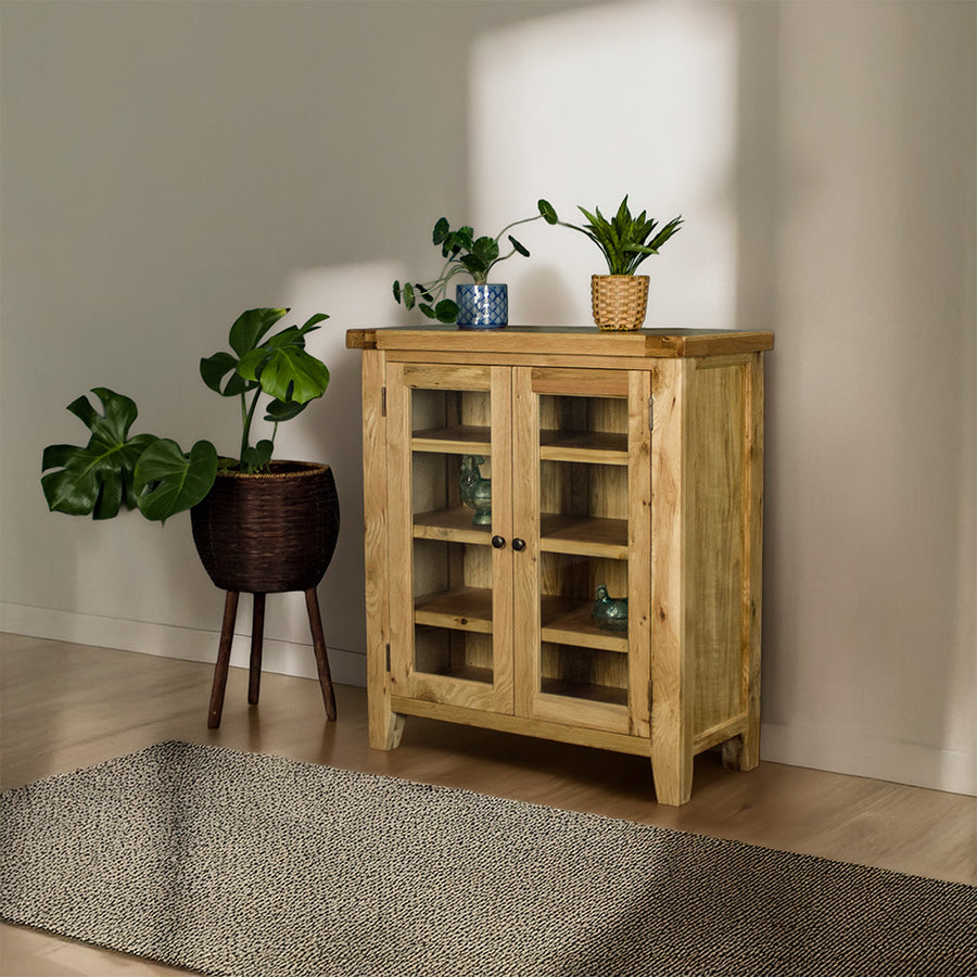 The front of the Yes Oak Display Cabinet. There are two blue glass ornaments on the middle shelves and two potted plants on top. There is a free standing potted plant next to the cabinet.