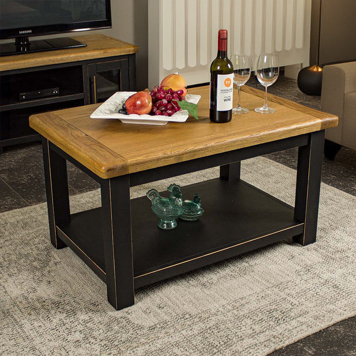 The front of the Cascais Oak-Top Coffee Table. There is a platter of fruit, a bottle of wine and two wine glasses on top. There are two blue glass ornaments on the shelf below.