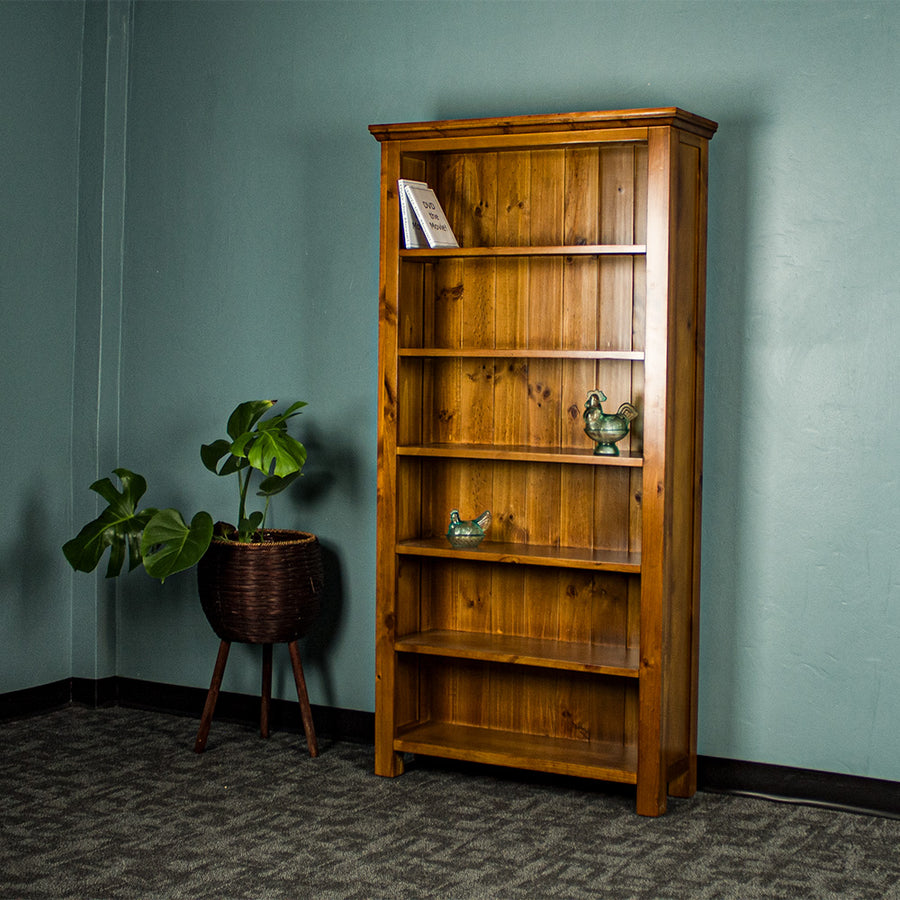 The front of the Montreal Large Pine Bookcase. There are two DVDs on the top shelf, and two blue glass ornaments on the middle two shelves. There is a free standing potted plant next to the bookshelf.