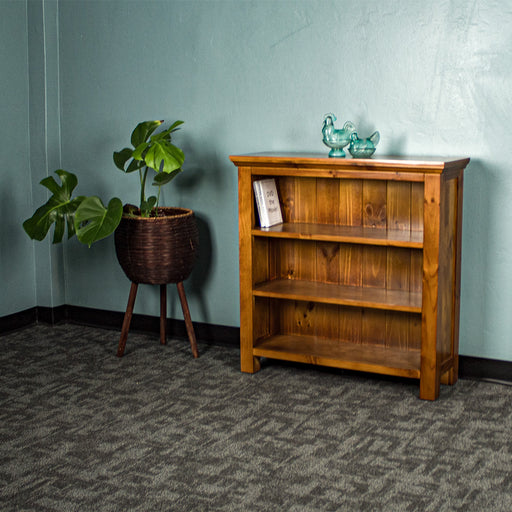 The front of the Montreal Short Pine Bookcase. There are two blue glass ornaments on top and two DVD cases on the top shelf. There is a free standing potted plant next to the bookshelf.