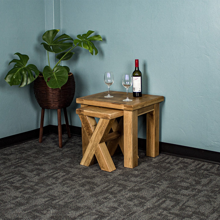 An overall view of the Amstel Oak Nesting Tables, with the smaller one sticking out slightly. There is a free standing potted plant next to the tables. There are two wine glasses and a bottle on top.