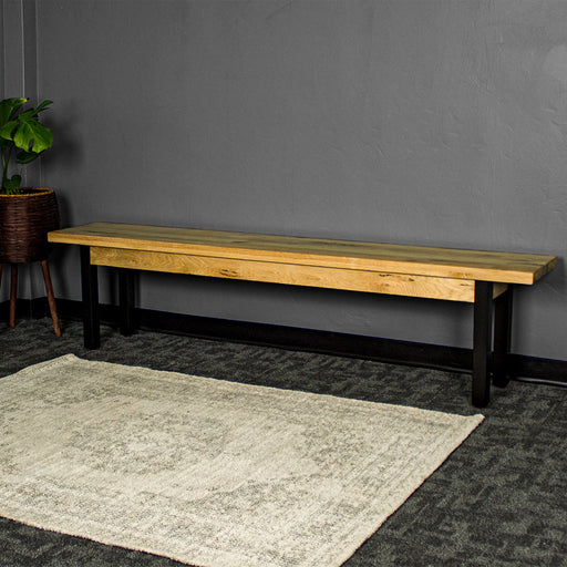 The front of the Golden Gate Long Oak Bench. There is a large gray rug in front and a potted plant next to the bench.