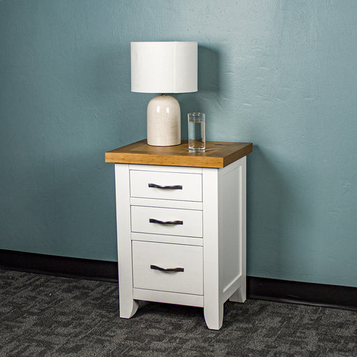 The front of the Felixstowe Pine Bedside Cabinet (White). There is a lamp and a glass of water on top.