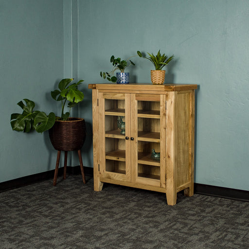 The front of the Yes Oak Display Cabinet. There are two blue glass ornaments on the middle shelves and two potted plants on top. There is a free standing potted plant next to the cabinet.