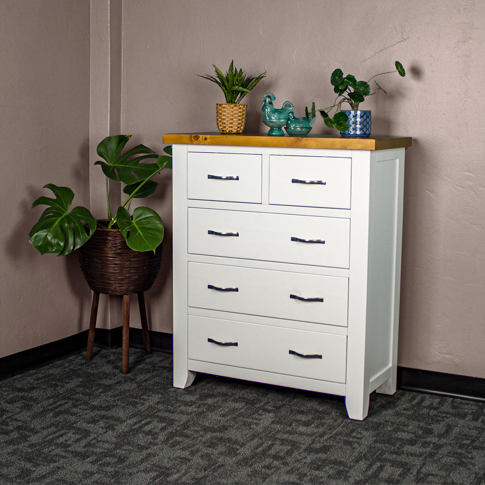 The front of the Felixstowe 5 Drawer Pine Tallboy. There are two potted plants on top with two blue glass ornaments in between. There is a free standing potted plant next to the tallboy.