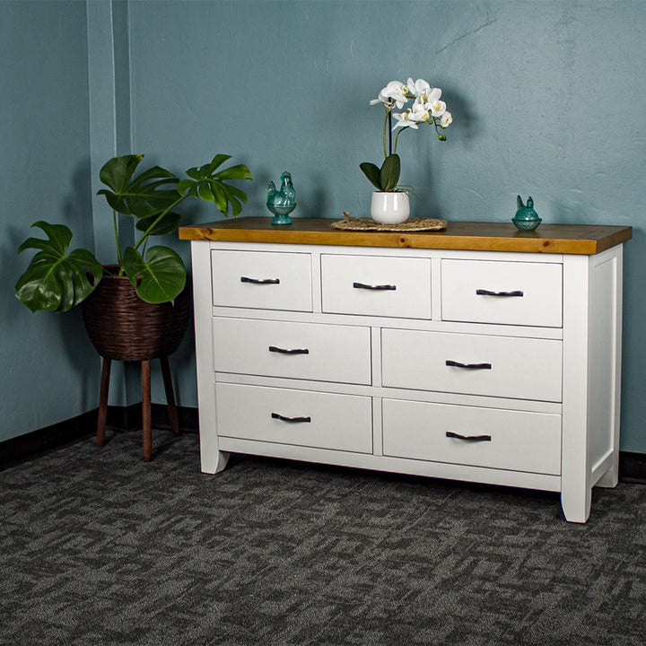 The front of the Felixstowe 7 Drawer Pine Lowboy. There are two blue glass ornaments on top with a small pot of white flowers on a rattan leaf in the middle of them. There is a free standing potted plant next to the lowboy.