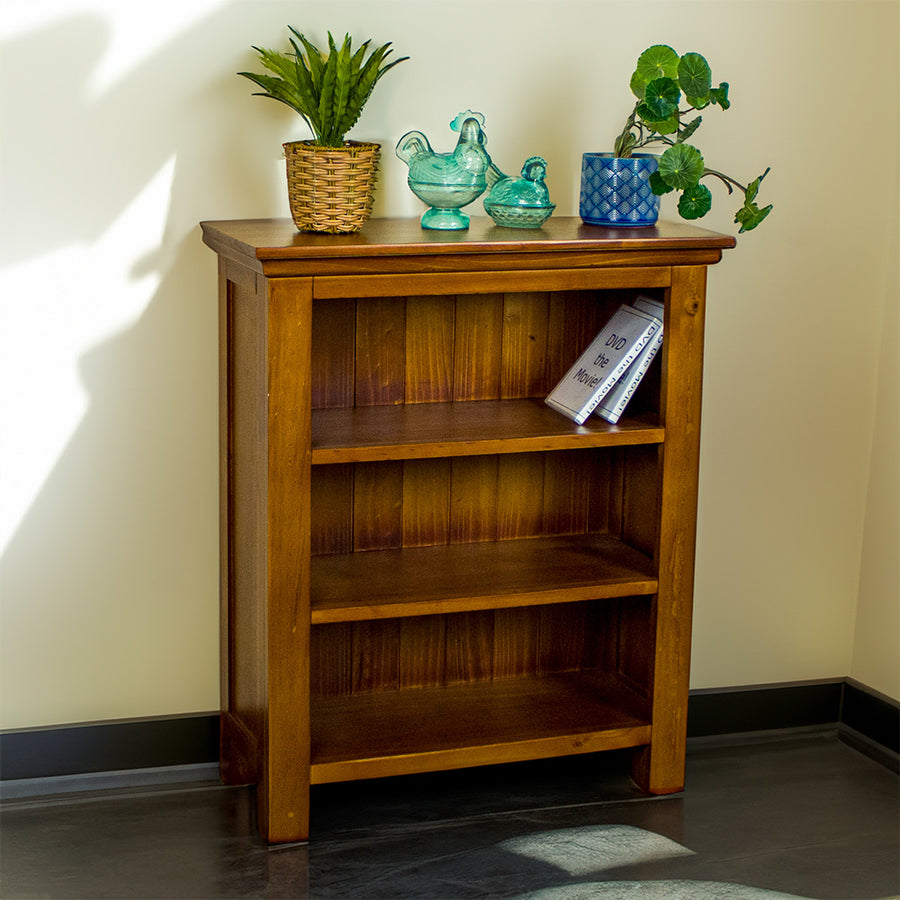 The front of the Montreal Low Pine Bookcase. There are two potted plants on top with two blue glass ornaments in between. There are two DVDs on the top shelf.
