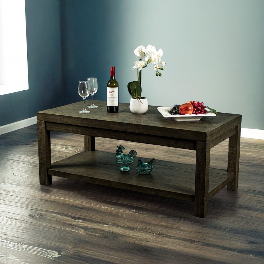 Front view of the brown Stonemill Recycled Pine Coffee Table. There are two wine glasses, a glass bottle, a potted plant and a platter of fruit on the top. There are two blue glass ornaments on the bottom shelf. There is also text in the bottom right corner that says "100% solid recycled pine" and "Environmentally friendly"
