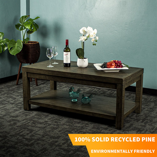 Front view of the brown Stonemill Recycled Pine Coffee Table. There are two wine glasses, a glass bottle, a potted plant and a platter of fruit on the top. There are two blue glass ornaments on the bottom shelf. There is also text in the bottom right corner that says "100% solid recycled pine" and "Environmentally friendly"