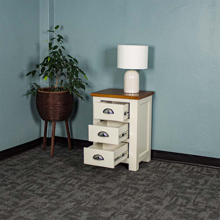 The Alton 3 Drawer Pine Bedside Cabinet with a Rimu stained top has its drawers open. There is a lamp on the top. It is next to a free standing potted plant.