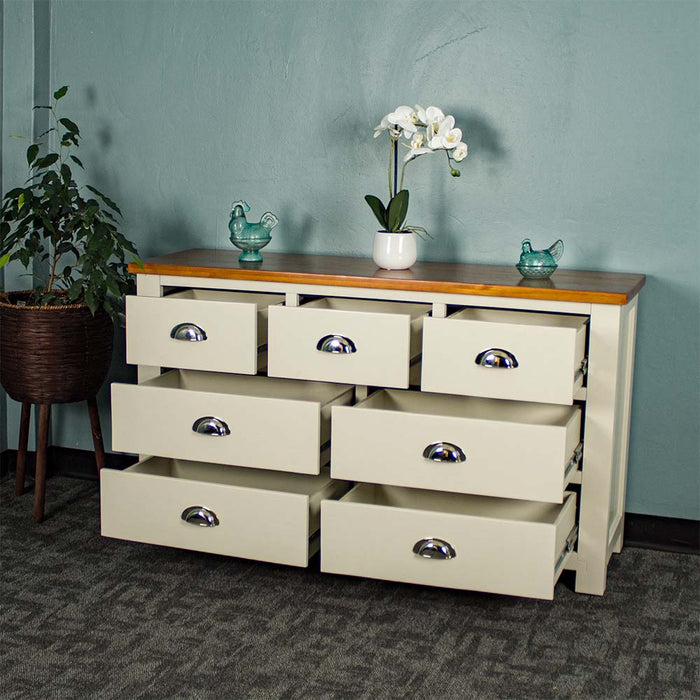 The front of the Alton 7 Drawer Pine Lowboy with its drawers open. There are two blue glass ornaments on top with a pot of white flowers in between. There is a free standing potted plant next to the lowboy.