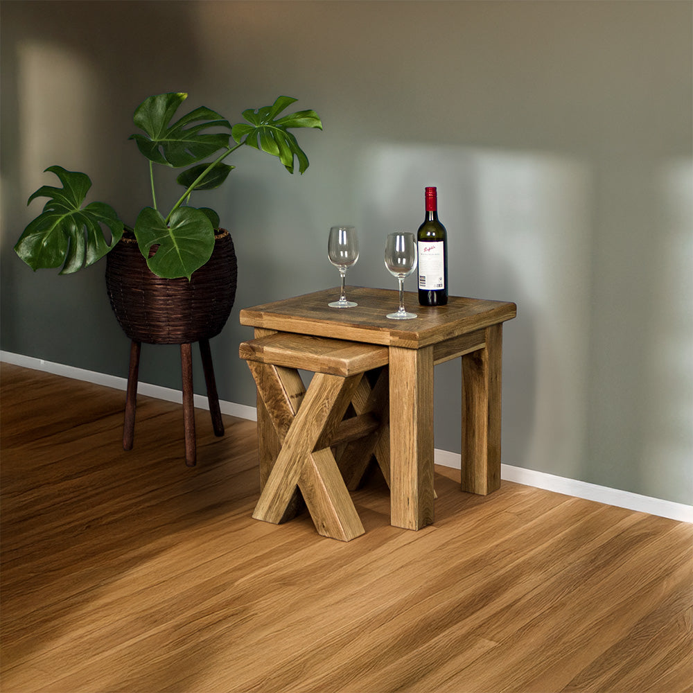 An overall view of the Amstel Oak Nesting Tables, with the smaller one sticking out slightly. There is a free standing potted plant next to the tables. There are two wine glasses and a bottle on top.