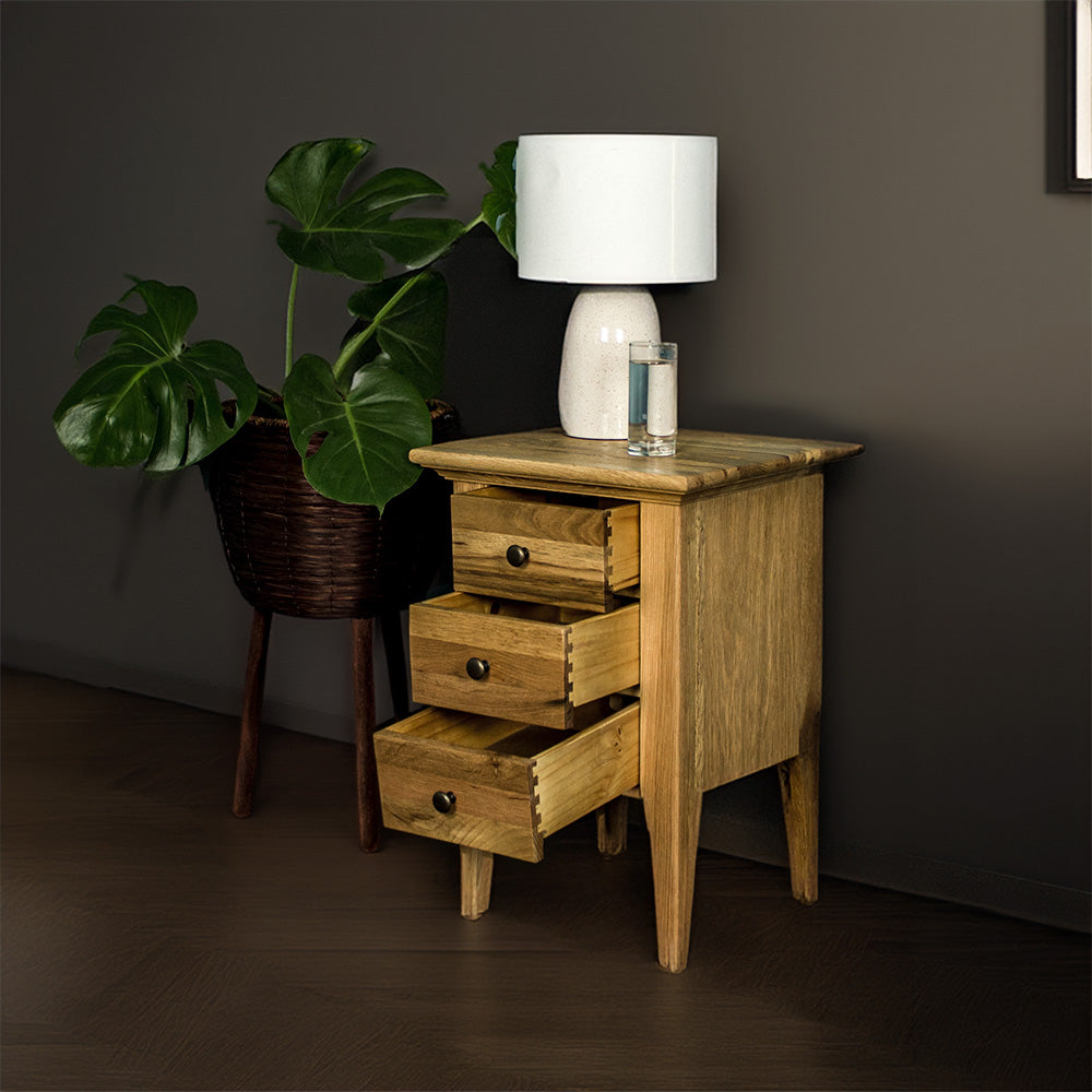 The front of the Beethoven Oak Bedside Table with its 3 Drawers open. There is a free standing potted plant next to it. There is a lamp and a glass of water on top.