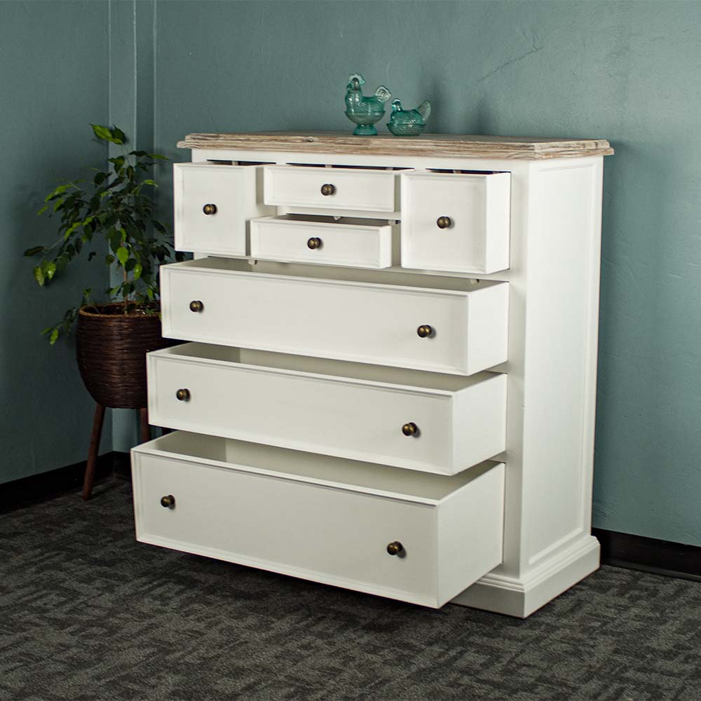 The front of the Biarritz 7 Drawer Tallboy / Chest of Drawer with its drawers open. There are two blue glass ornaments on top and a free standing potted plant next to it.