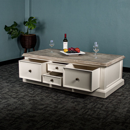 An overall view of the Biarritz 4 Drawer Extra-Large Coffee Table with its drawers open. There are three wine glasses, a bottle and a platter of fruit on top. There is a free standing potted plant in the background.