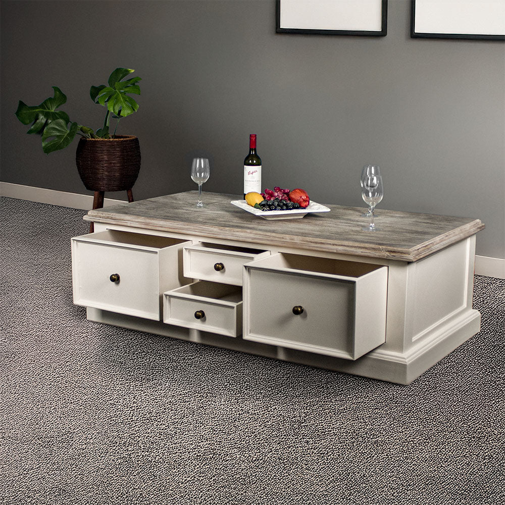 An overall view of the Biarritz 4 Drawer Extra-Large Coffee Table with its drawers open. There are three wine glasses, a bottle and a platter of fruit on top. There is a free standing potted plant in the background.