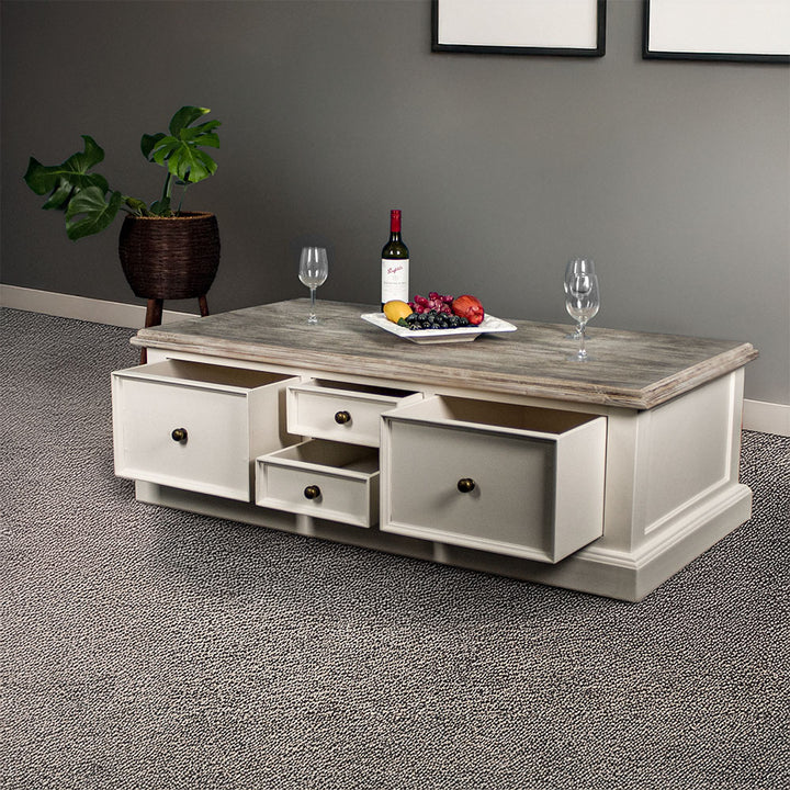 An overall view of the Biarritz 4 Drawer Extra-Large Coffee Table with its drawers open. There are three wine glasses, a bottle and a platter of fruit on top. There is a free standing potted plant in the background.