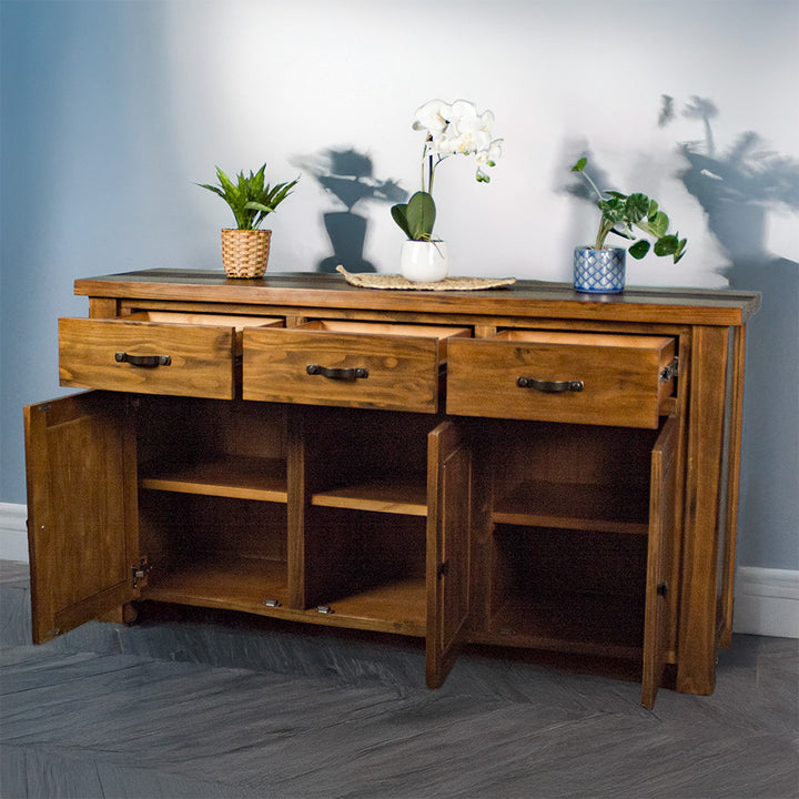 The front of the Botanica 3-Drawer Large Buffet, with its drawers and doors open. There are three potted plants on top, with the middle one on top of a rattan leaf.