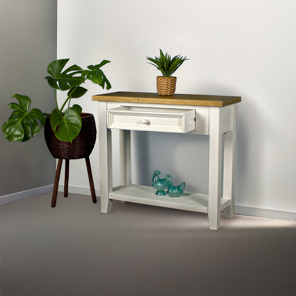 Front view of the Byron Recycled Pine Hall Table with its drawer open. There is a standing potted plant to the side, a small potted plant on the top, and two blue glass ornaments on the lower shelf.