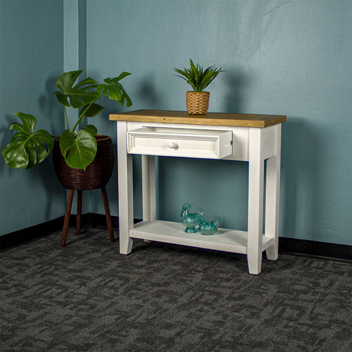 Front view of the Byron Recycled Pine Hall Table with its drawer open. There is a standing potted plant to the side, a small potted plant on the top, and two blue glass ornaments on the lower shelf.