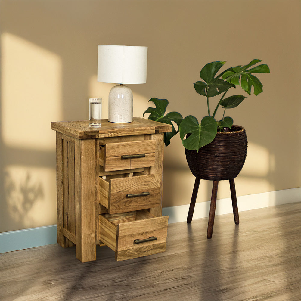 An overall view of the Camden 3 Drawer White Oak Bedside Table with its drawers open. There is a glass of water and a lamp on top. There is a free standing potted plant next to the bedside table.