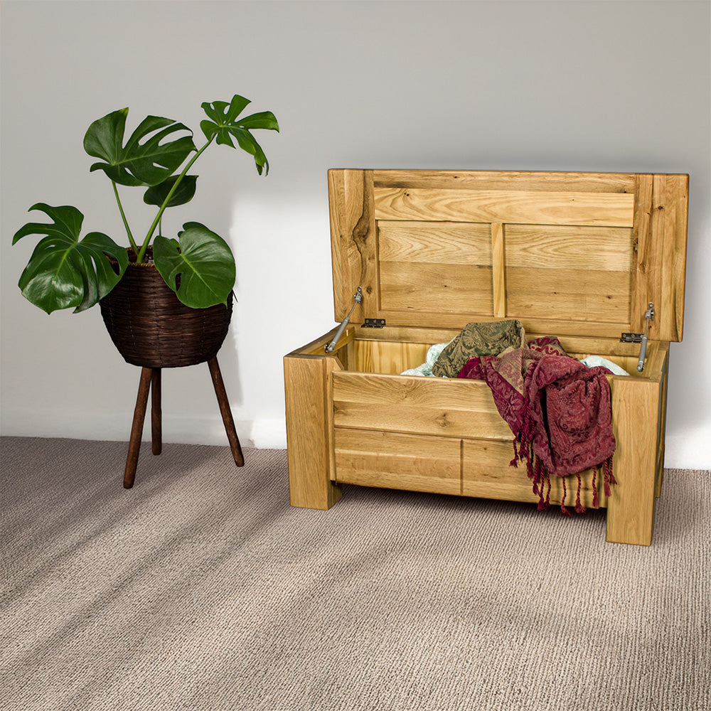 An overall view of the Camden Oak Blanket Box, which is open with blankets hanging out of it. There is a free standing potted plant next to it.