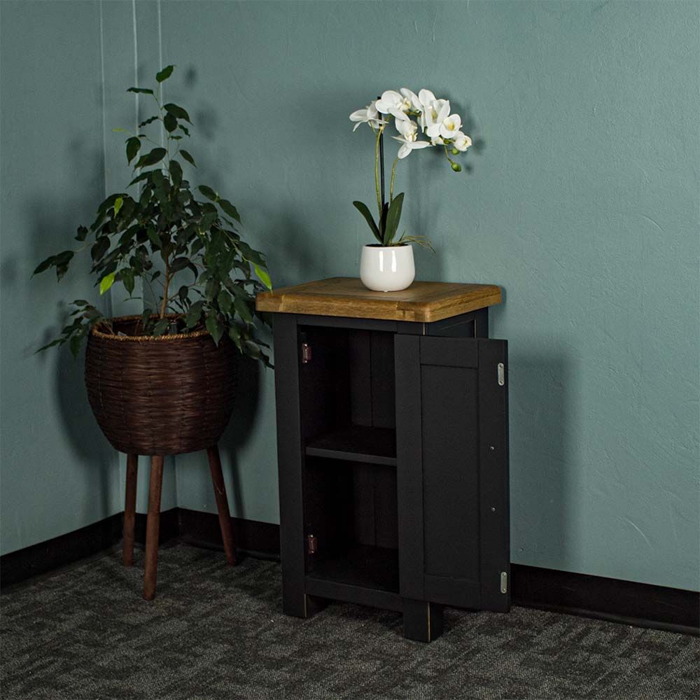 The front of the Cascais Small Black Cupboard with its door open. There is a small pot of white flowers on top. There is a free standing potted plant next to the cupboard.