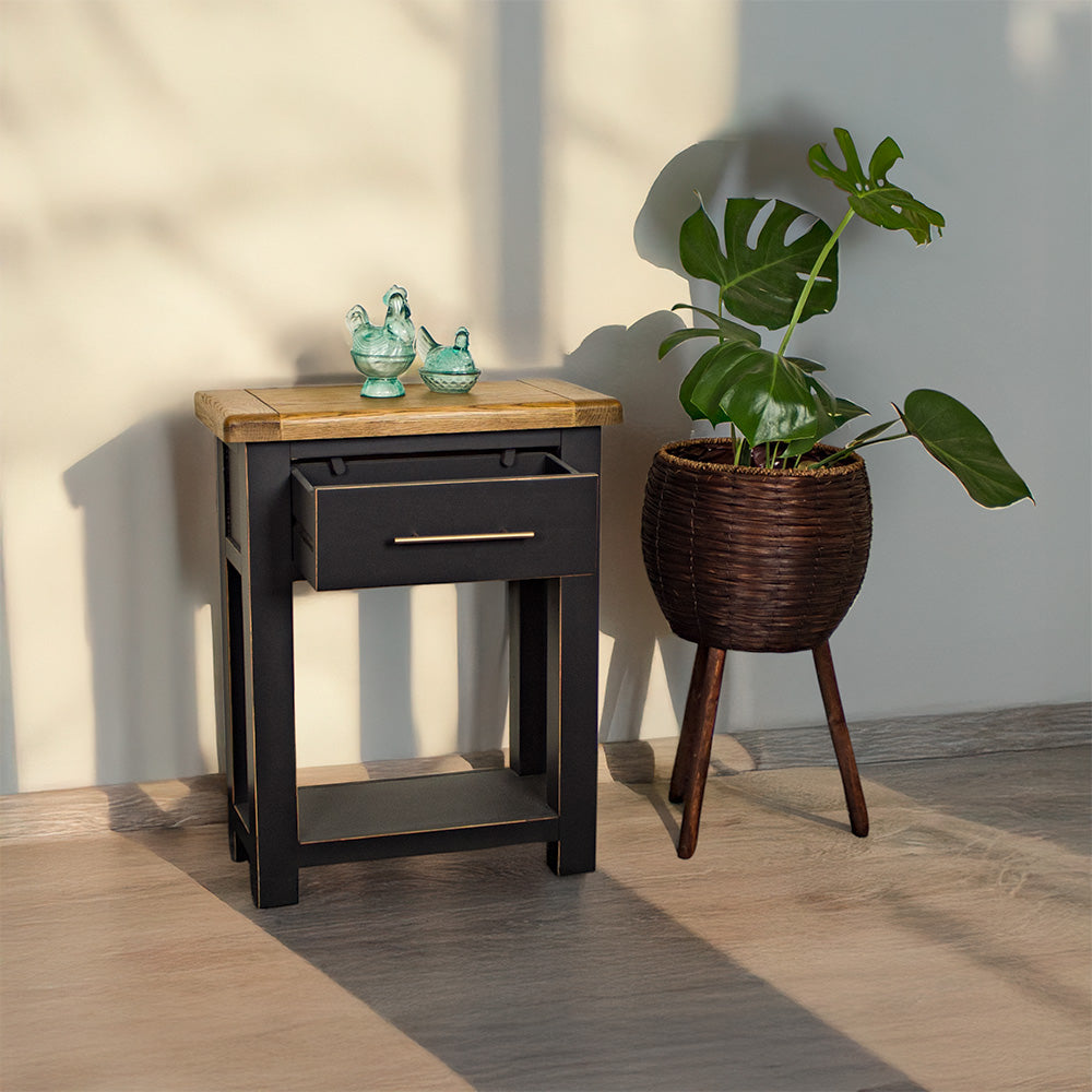 An overall view of the Cascais Black Hall Table, with its drawer open. There are two blue glass ornaments on top with a tall, free standing potted plant next to it.