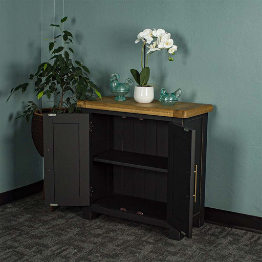 The front of the Cascais Oak-Top Small Sideboard with its doors open. There are two blue glass ornaments on top with a small pot of white flowers in between. There is a free standing potted plant next to the unit.